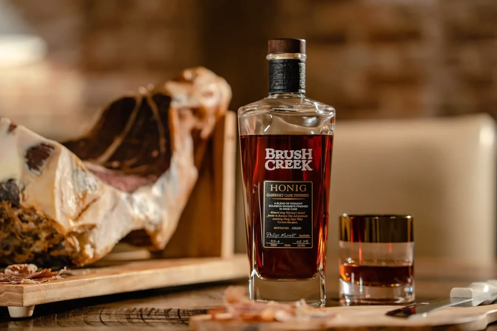 Bottle of Brush Creek Honig Cabernet Cask Finished bourbon whiskey sitting on table next to glass with the bourbon whiskey poured into it